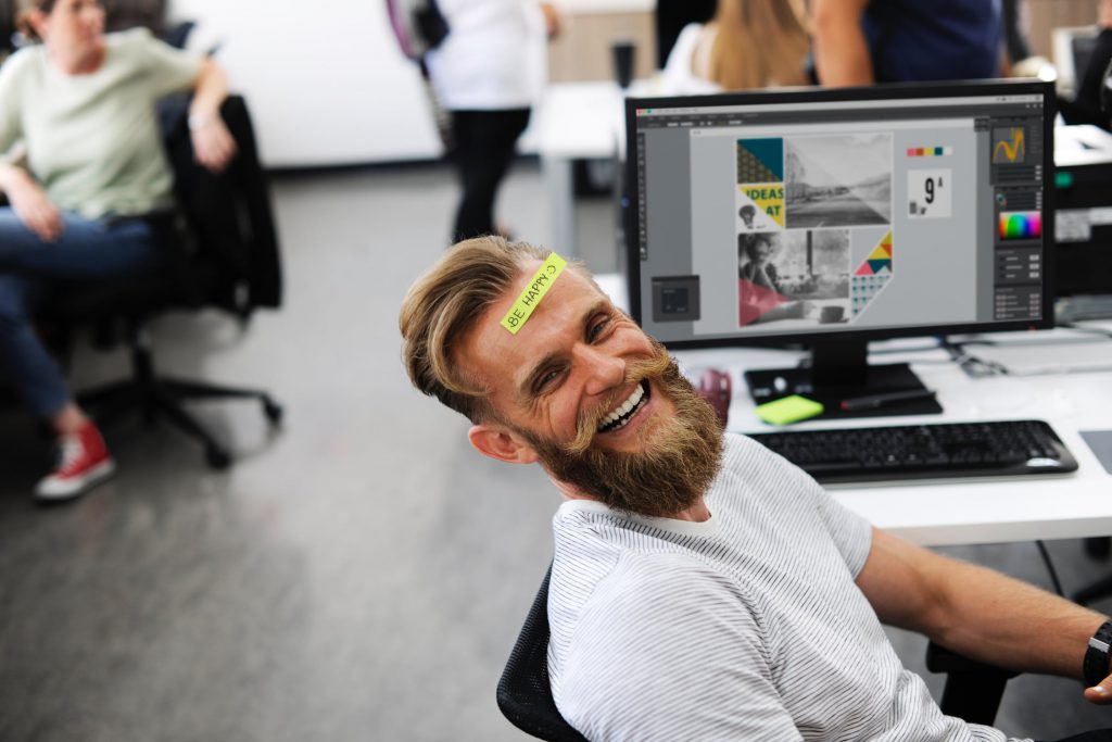 A graphic design employee at his desk with a post it note on his head that says "be happy" | Work Place Culture HR Consulting