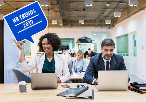 Employees in a modern decorated office holding a sign that says HR Trends 2019